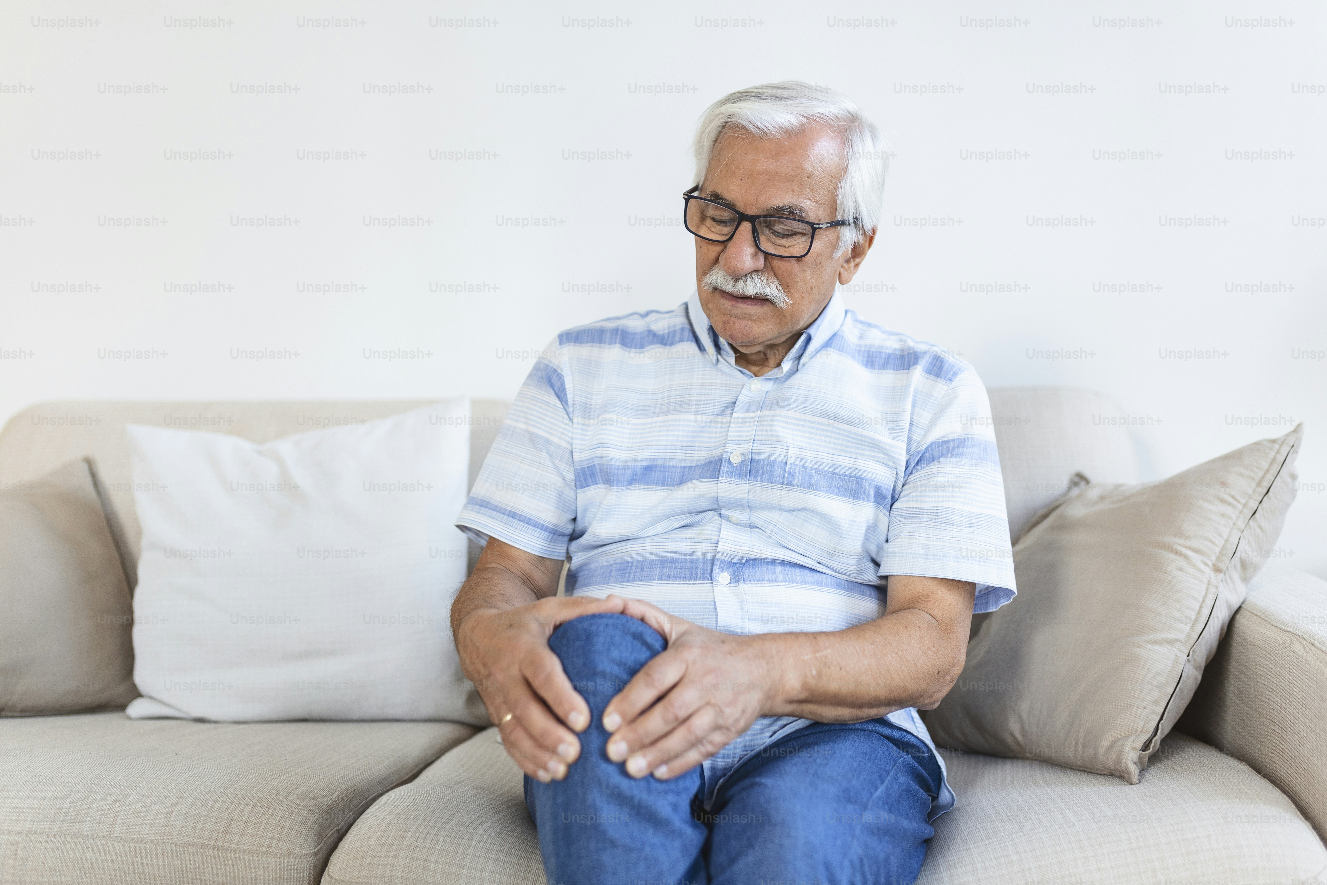 Elderly man sitting on a sofa at home and touching his painful knee. people, health care and problem concept - unhappy senior man suffering from knee ache at home
