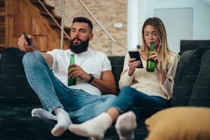 Young beautiful couple drinking beer at home and using a smartphone while relaxing on the couch and watching television on the weekend