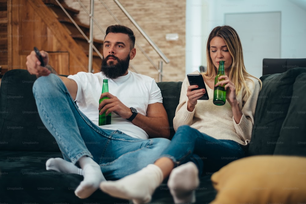 Young beautiful couple drinking beer at home and using a smartphone while relaxing on the couch and watching television on the weekend