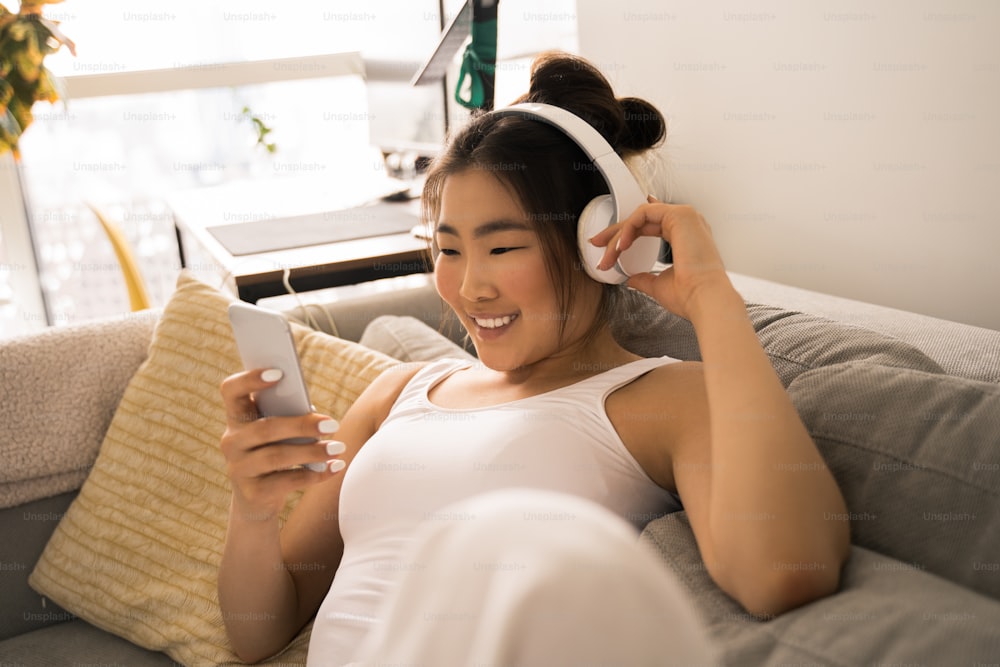 Close up of calm young woman in wireless headphones relaxing on sofa at home and dreaming while listen to music. Happy asian girl looking at the smartphone screen with pleasure smile