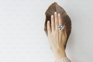Elegante anillo redondo moderno en hermosa mano sobre fondo de hoja grande seca marrón en pared blanca. Anillo de vidrio fundido de moda inusual en la mano femenina con manicura blanca. Concepto de cuidado