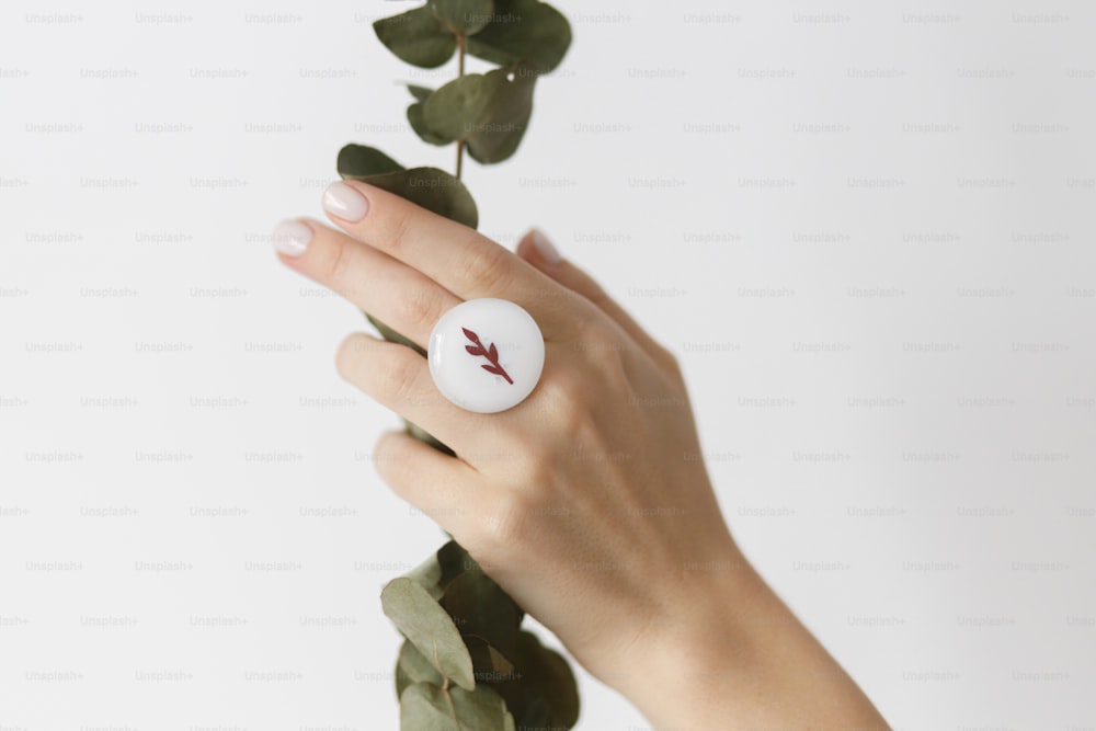 Elegante anello rotondo bianco moderno su bella mano con ramo di eucalipto su sfondo bianco. Insolito anello in vetro fuso alla moda su mano femminile con manicure bianca. Concetto ecologico e di cura
