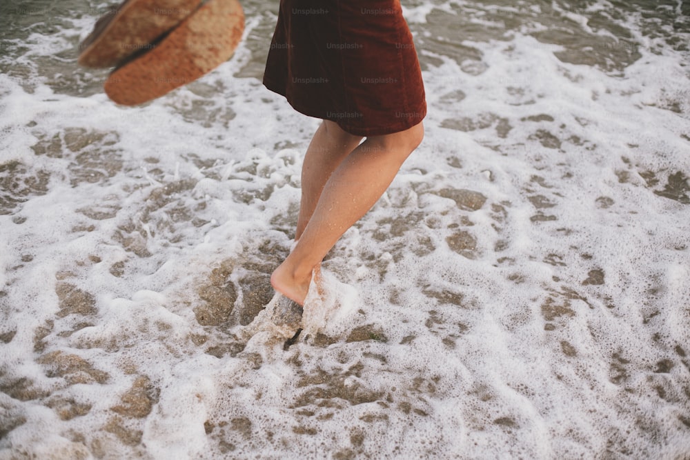 Unbeschwerte Hipster-Frau, die barfuß in Meereswellen am Strand läuft und Flip-Flops in der Hand hält. Ruhiger entspannender Moment. Füße im Wasser mit Schaum, Sommerurlaub. Achtsamkeit