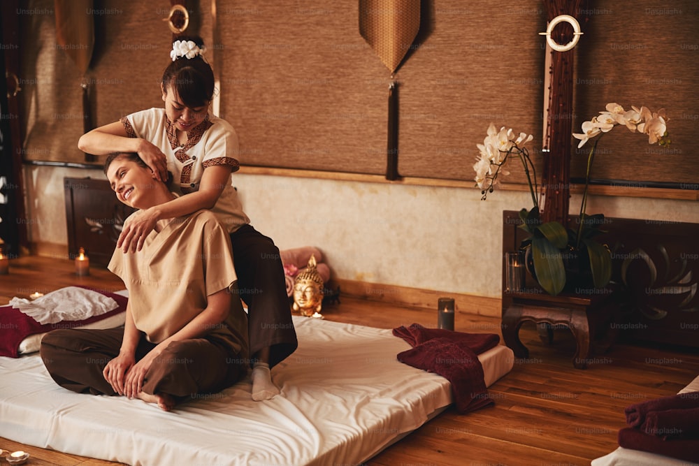 Experienced Asian massage specialist standing behind sitting woman in lotus position and pressing her palm to client ear