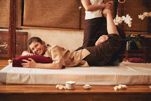 Visitante satisfeita do salão de massagem deitada de barriga para baixo no colchão e sorrindo com massagista esfregando o pé esquerdo