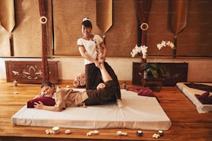 Female resting on mattress with pillow, lying on wooden floor, while masseuse in national outfit performing her leg stretching