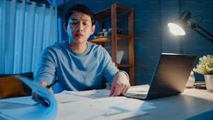 아시아 프리랜서 사업가는 밤에 집에서 초과 근무를 하고 집에서 거실의 책상에 있는 서류 작업 차트로 가득 찬 노트북 컴퓨터에 초점을 맞추고 COVID-19 전염병 개념 동안 집에서 일합니다.