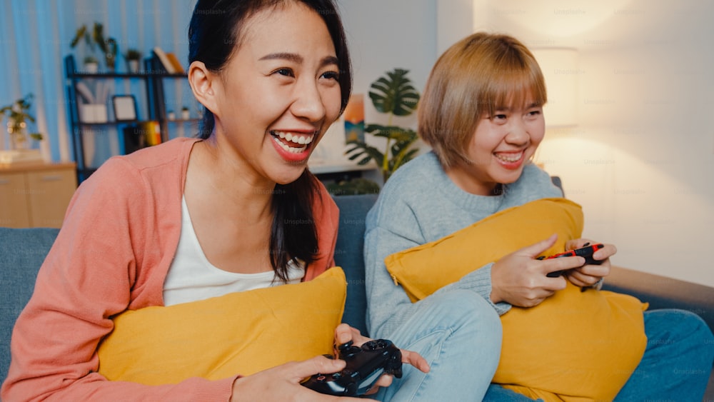 Lesbian LGBTQ women couple play video game at home. Young Asia lady using wireless controller having funny happy moment on sofa in living room at night. They have great and fun time celebrate holiday.
