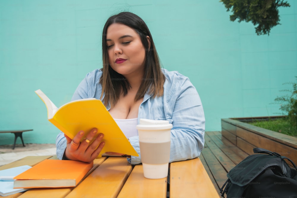 Portrait of young plus size woman enjoying free time and reading a book while sitting outdoors at coffee shop.