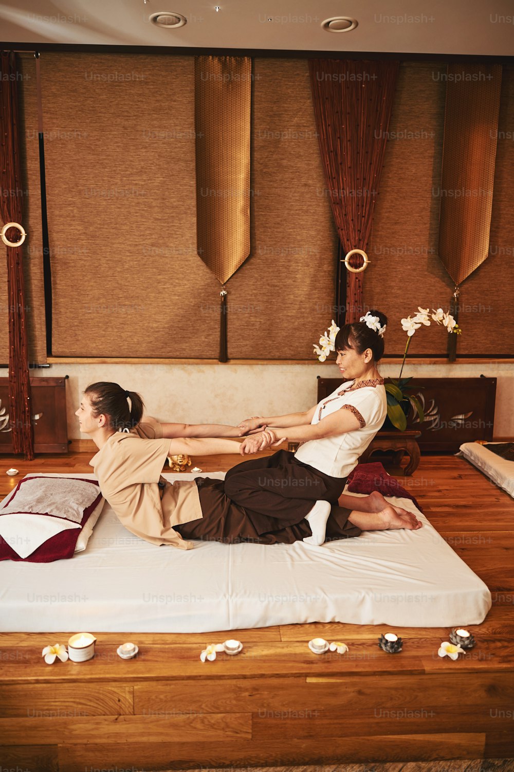 Woman Thai massage professional sitting with knees on female butt and pulling her by hands on mattress