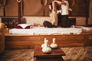 Herbal balls standing on wooden chair in spa salon with two people performing Thai massage in the background