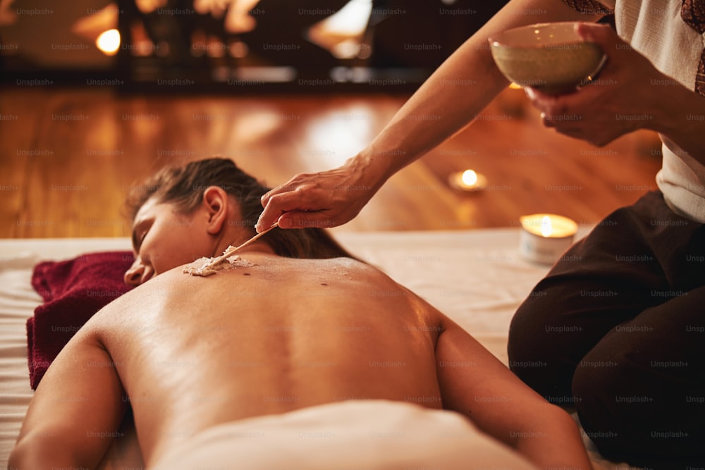 Masseuse raising clay bowl in left hand while making use of wooden stick for distributing massage cream on woman back