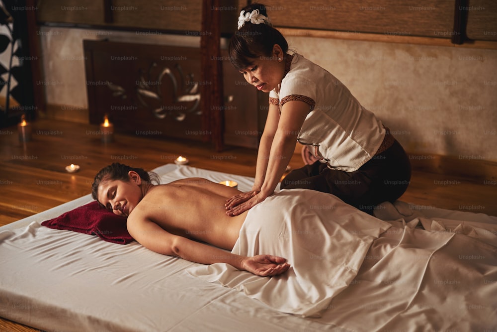Female sitting on her knees over relaxing client and treating her naked lower back with Thai massage