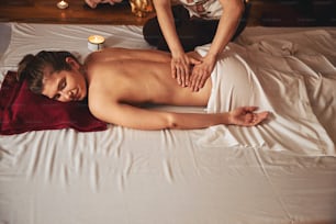 Femme aux yeux fermés allongée sur un matelas avec une bougie près de son côté pendant qu’une spécialiste de la beauté fait un massage de ses reins
