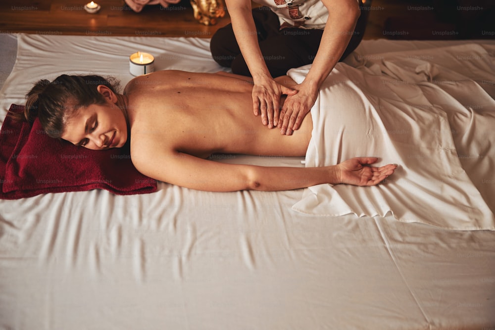 Woman with closed eyes lying on mattress with candle near her side while beauty specialist doing massage of her loin