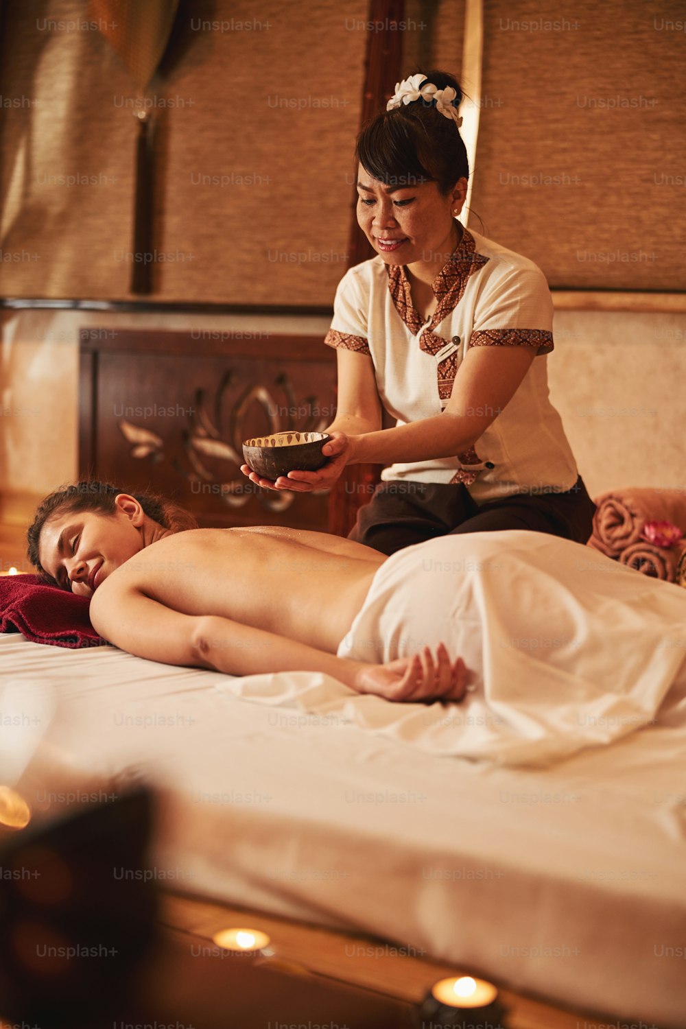 Careful spa salon worker stretching her arms over lying woman while holding clay bowl with oil for massage