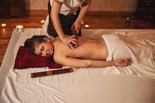 Lady with naked back lying face-down on mat on the floor while massagist tending her back with roller