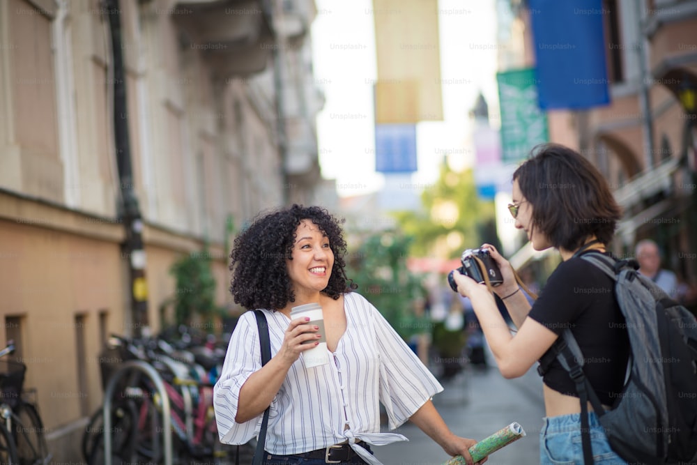 市内の観光客。 コーヒーを飲みながら通りに立つ笑顔の女性。友人の写真を撮る女性。焦点は背景にあります。