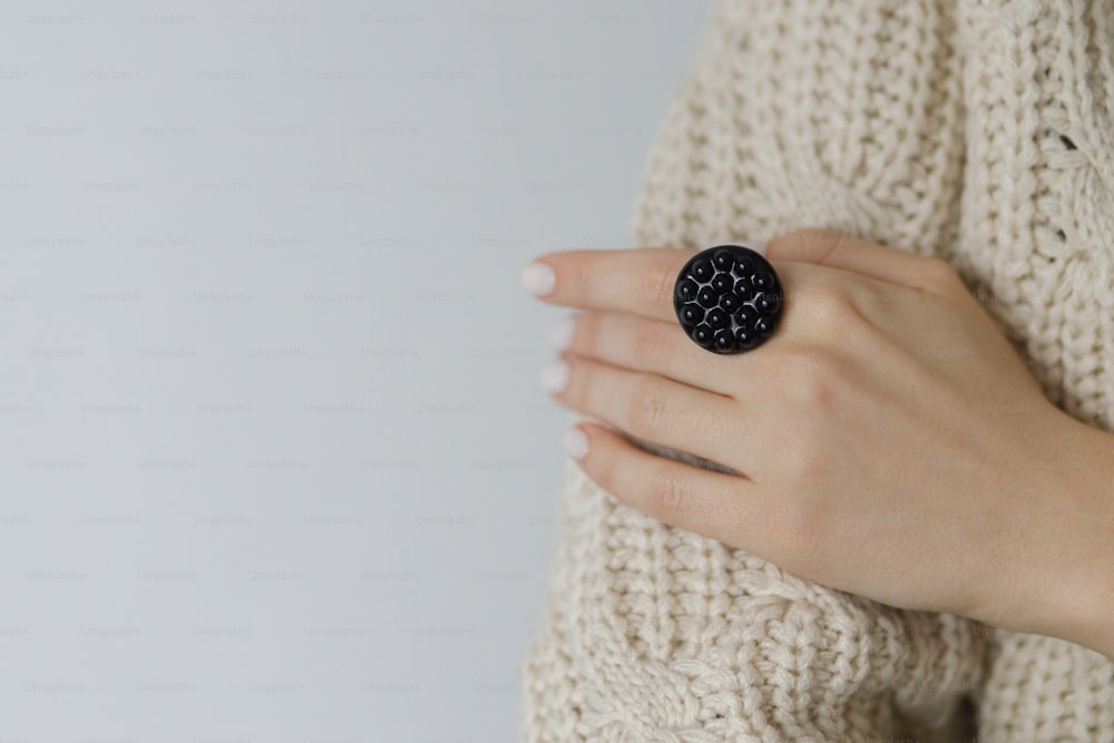 Stylish modern round black ring on beautiful hand on background beige sweater and white wall. Fashionable female in sweater with unusual fused glass accessory. Contemporary gift