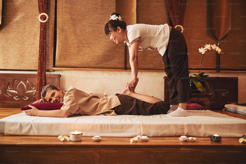 Female standing with legs on floor mattress and bending over lying woman while kneading her feet during Thai massage