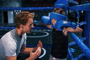 Young boxing coach is helping little boy in protective wear on the ring between the rounds.