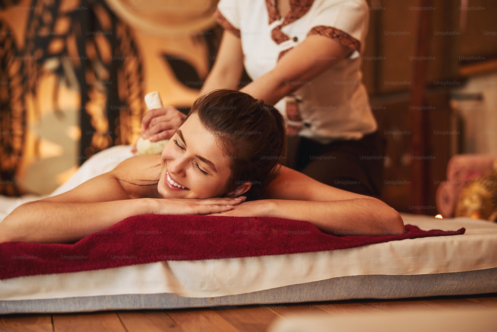 Donna felice che riposa il viso sulle mani giunte mentre il membro del personale della spa le tratta la schiena con un massaggio Impiastro a base di erbe tailandese