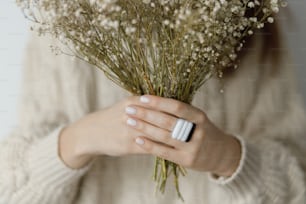 Hermosa mujer con estilo con anillo cuadrado moderno sosteniendo flores secas, de cerca. Mujer de moda en suéter con accesorios inusuales de vidrio fundido y manicura blanca. Concepto de belleza y cuidado