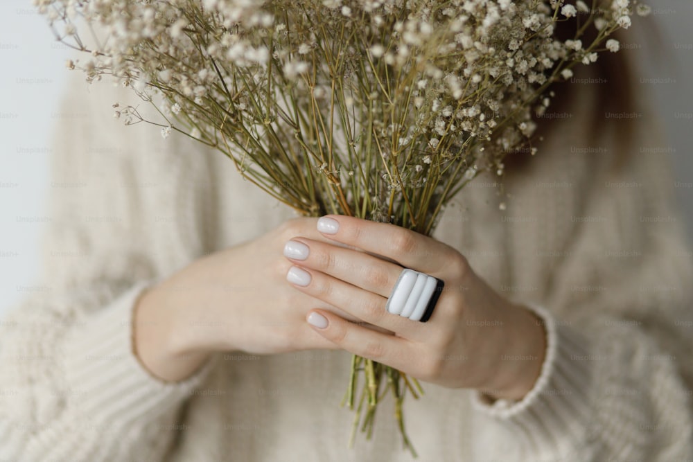Beautiful stylish woman with modern square ring holding dried flowers, close up. Fashionable female in sweater with unusual fused glass accessories and white manicure. Beauty and care concept