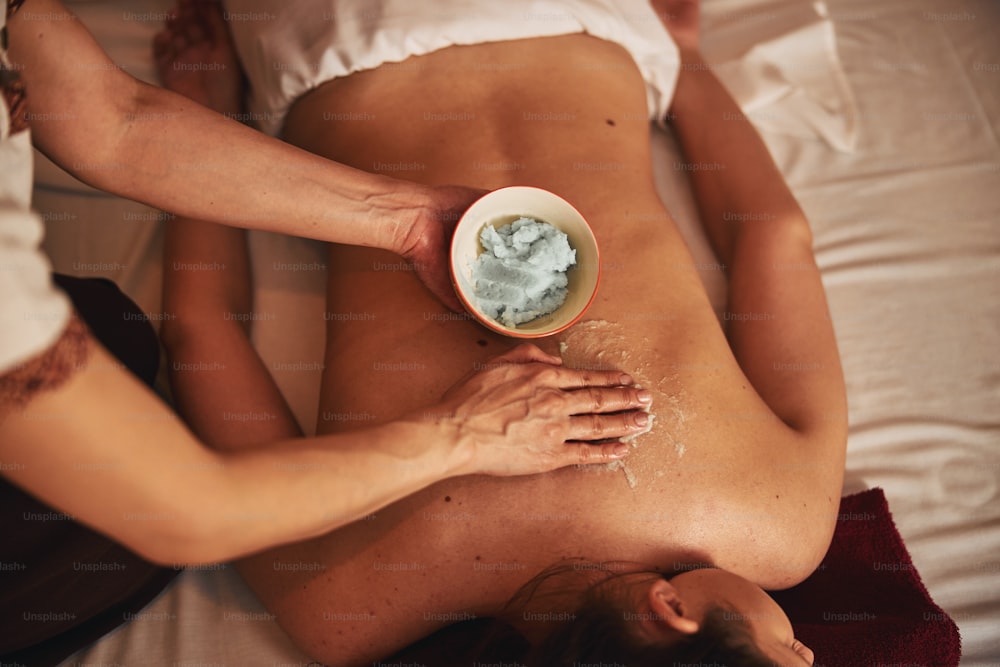 Spa worker using beauty balm from bowl to prepare woman back for massage treatment