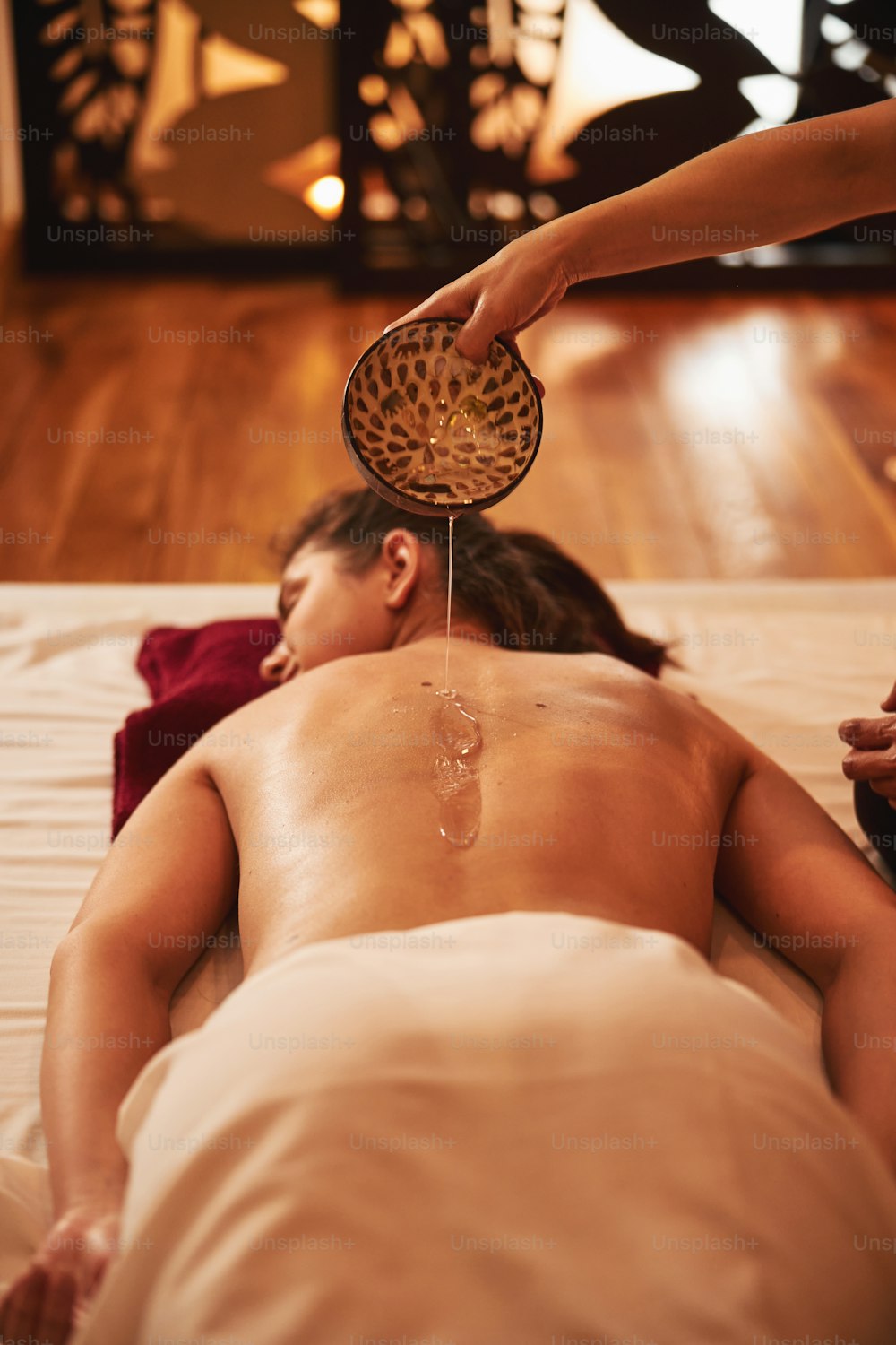 Hand of masseuse turning dish with massage oil over body of woman, lying on floor bed in spa salon