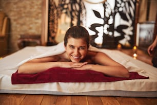 Cheerful lady laying her chin on folded hands while resting on mattress on the wooden floor of spa salon