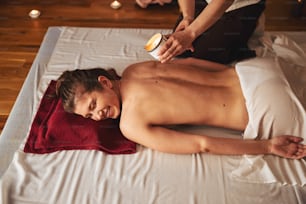 Female on white mat giggling while person spilling hot wax on her bare back skin from massage candle