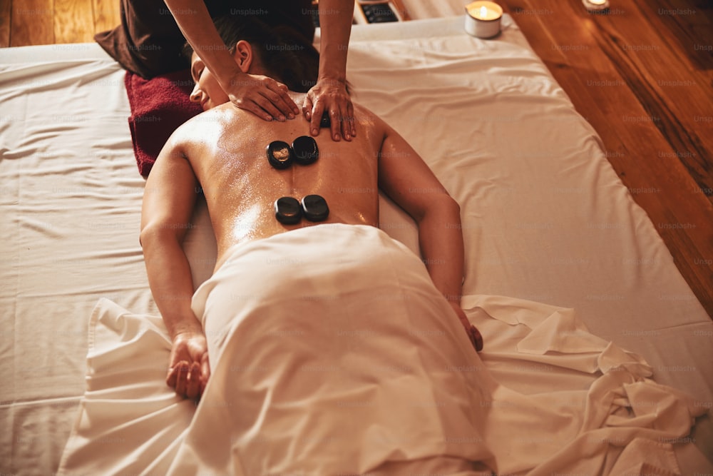 Massaggiatore che strofina la parte superiore della schiena della donna con una pietra nera in mano mentre altre quattro pietre calde si trovano sulla schiena