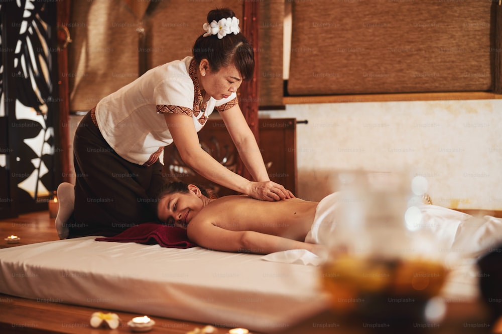 Mature Asian massage specialist working in spa and providing thermotherapy to young female with the help of stones