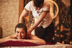 Customer of massage salon feeling joy and smiling to camera while Thai worker kneading her shoulders with both hands