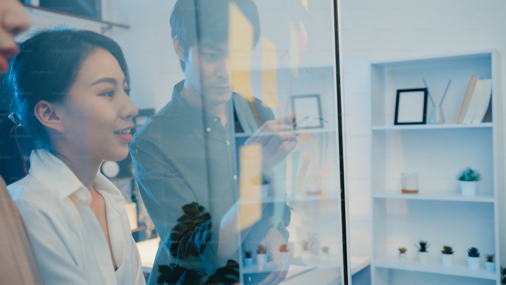 Asia businesspeople stand behind transparent glass wall listen manager pointing progress work and brainstorm meeting and worker post sticky note on wall. Business inspiration, Share business ideas.