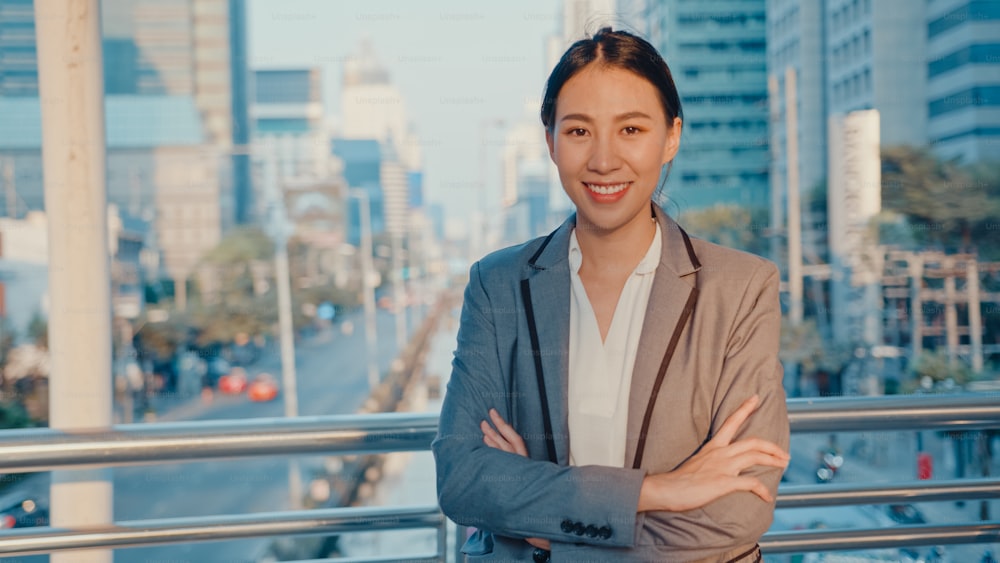 Successful young Asia businesswoman in fashion office clothes smiling and looking at camera while happy standing alone outdoors in urban modern city in the morning. Business on the go concept.