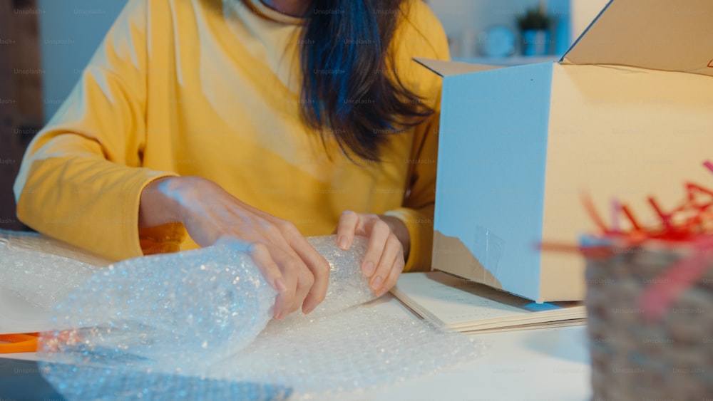 Junge asiatische Geschäftsfrau, die Glas verpackt, verwendet Luftpolsterfolie zum Verpacken von Stützschäden zerbrechliches Produkt im Home Office in der Nacht. Kleinunternehmer, Online-Marktlieferung, Lifestyle-Freelance-Konzept.