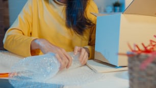 Young Asia businesswoman packing glass use bubble wrap for packing support damage fragile product in home office at night. Small business owner, online market delivery, lifestyle freelance concept.