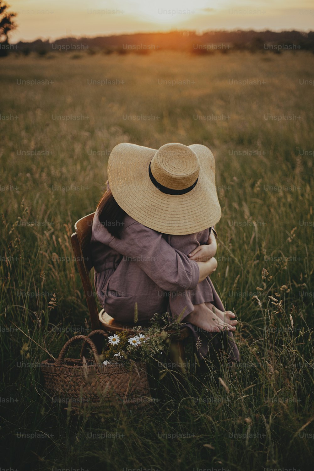 Beautiful woman in linen dress and straw hat sitting on rustic chair  in summer meadow. Young female relaxing with basket of flowers in sunset in countryside. Atmospheric tranquil moment