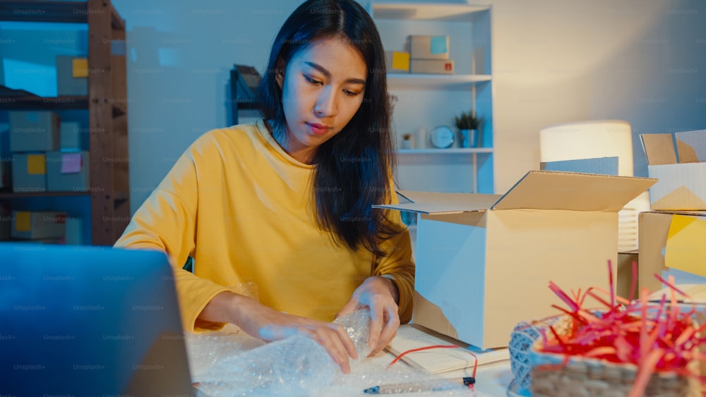 Young Asia businesswoman packing glass use bubble wrap for packing support damage fragile product in home office at night. Small business owner, online market delivery, lifestyle freelance concept.