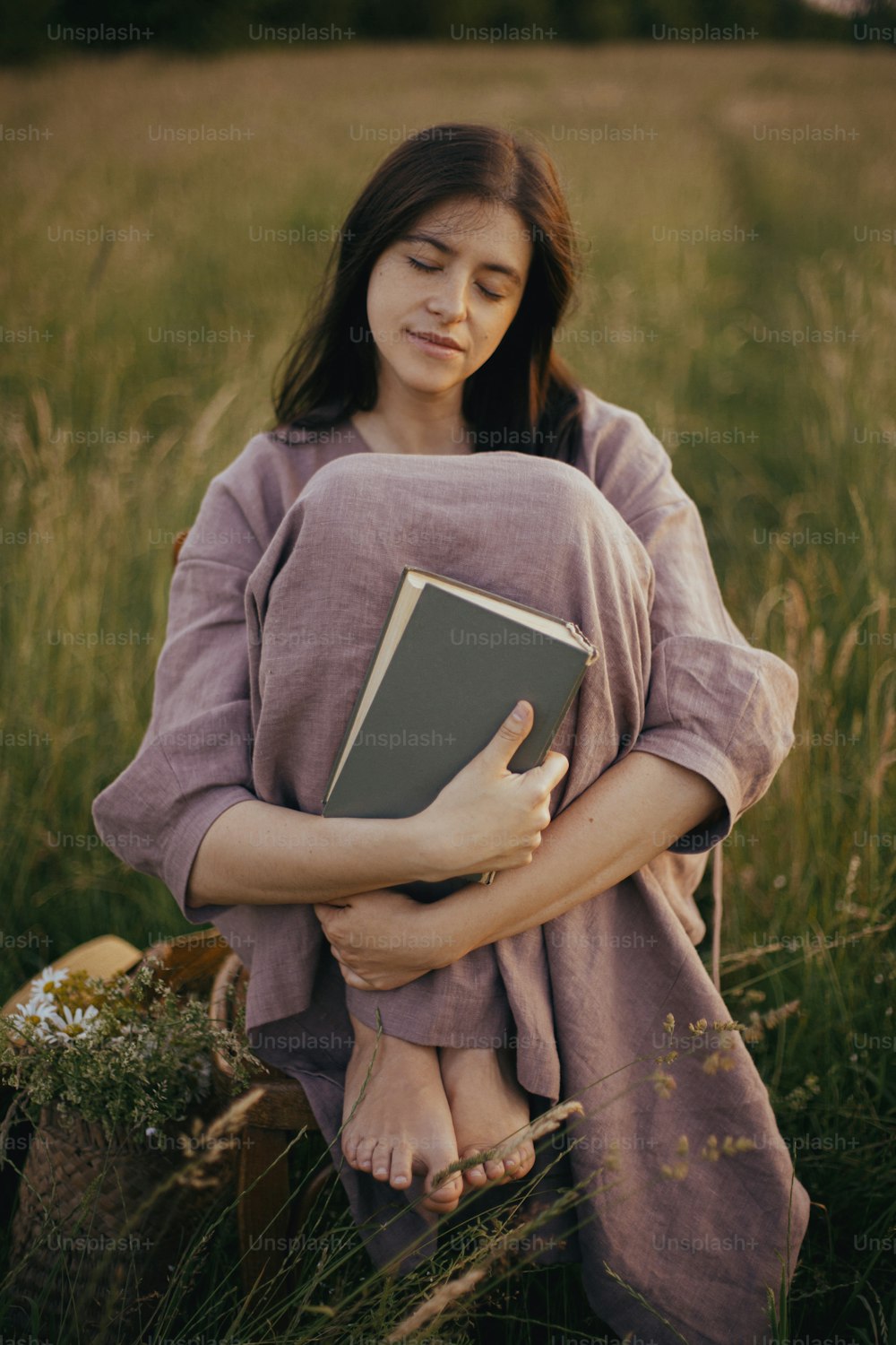 Hermosa mujer vestida de lino sentada en una silla rústica y soñando en la pradera de verano. Mujer joven relajante con libro y cesta de flores en el campo. Momento despreocupado atmosférico