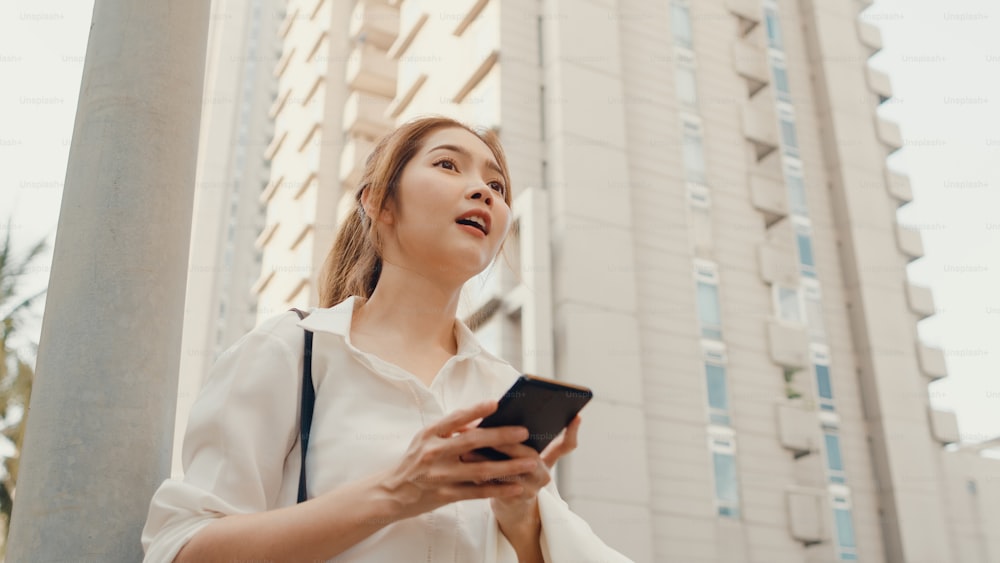패션 사무복을 입은 성공적인 젊은 아시아 사업가는 택시를 타고 도시 현대 도시의 야외에 서 있는 동안 스마트폰을 사용합니다. 이동 중 비즈니스 개념.