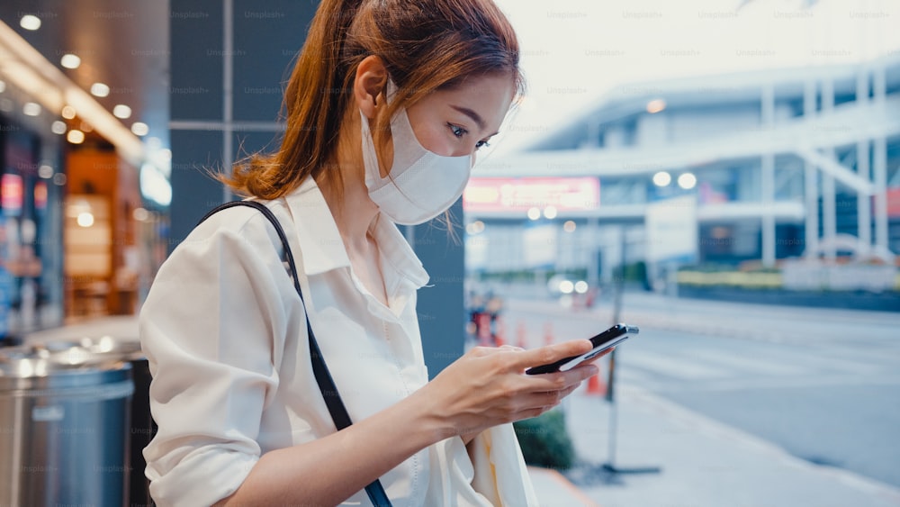 Young Asia businesswoman in fashion office clothes wearing medical face mask using smart phone typing text message while sitting outdoors in urban modern city at night. Business on the go concept.