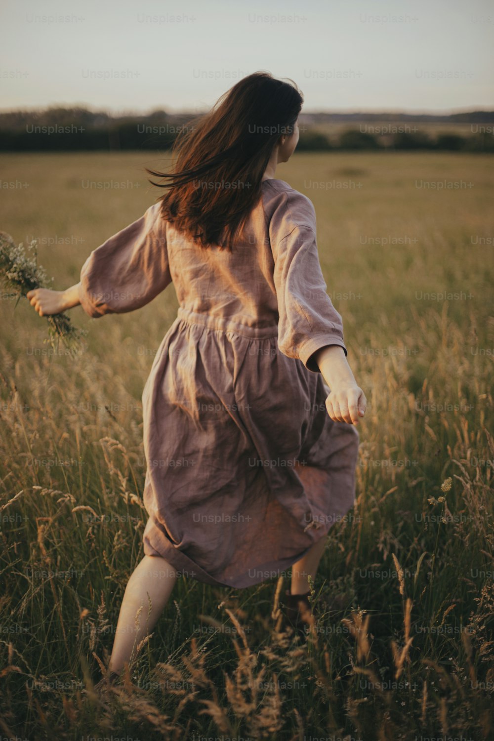 Beautiful woman in linen dress running with wildflowers in hand in summer meadow in sunset. Atmospheric carefree moment. Stylish young female in rustic dress enjoying free evening in countryside
