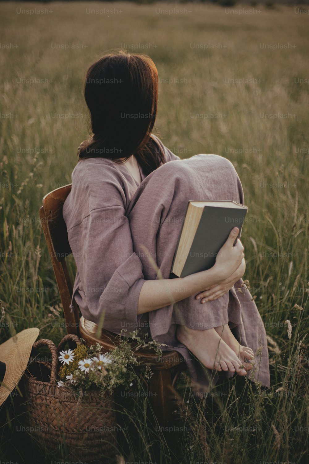 Mulher bonita no vestido de linho sentada na cadeira rústica e sonhando no prado de verão. Fêmea nova relaxando com livro e cesta de flores no pôr do sol do campo. Momento de tranquilidade atmosférica