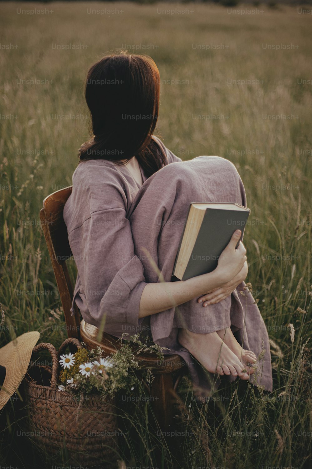 Beautiful woman in linen dress sitting on rustic chair and dreaming in summer meadow. Young female relaxing with book and basket of flowers in countryside sunset. Atmospheric tranquil moment