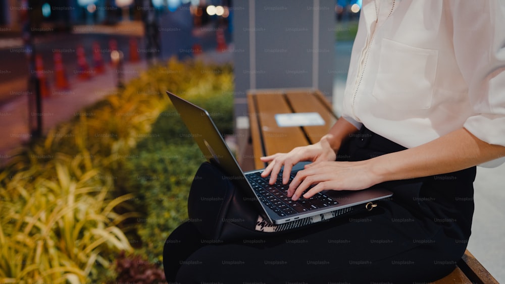 Successful young Asia businesswoman in fashion office clothes using laptop work remotely while sitting alone outdoors in urban modern city in the evening. Business on the go concept.