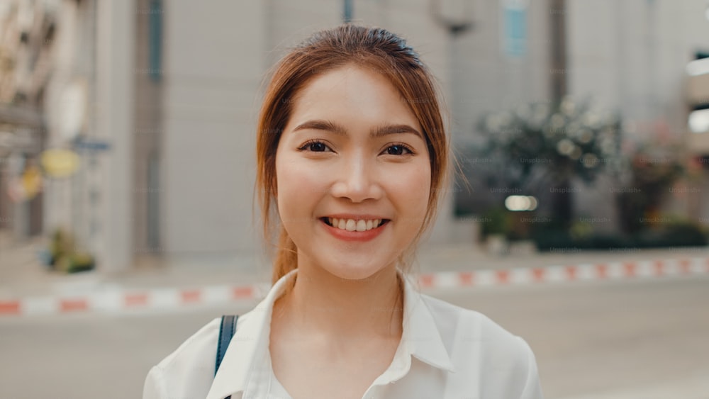 Erfolgreiche junge asiatische Geschäftsfrau in modischer Bürokleidung, die lächelt und in die Kamera schaut, während sie morgens glücklich ist, allein im Freien in der urbanen modernen Stadt zu stehen. Business on the go-Konzept.