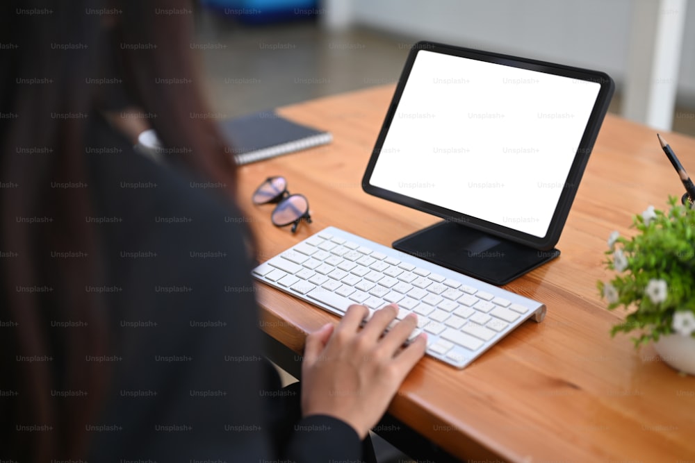Vista trasera de una mujer de negocios escribiendo en un teclado inalámbrico mientras trabaja con una tableta digital.